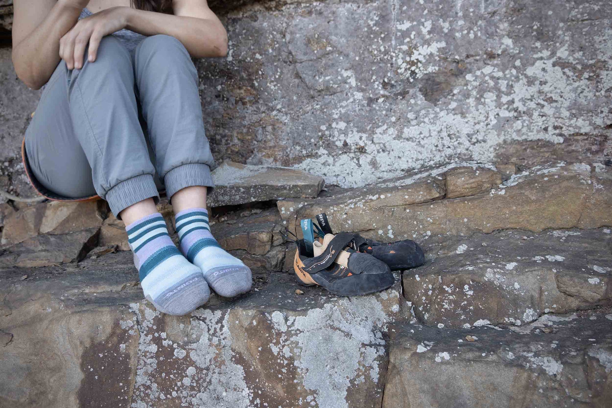 Climber sitting on ledge wearing colorful striped Point6 1/4 crew socks, with climbing shoes nearby.